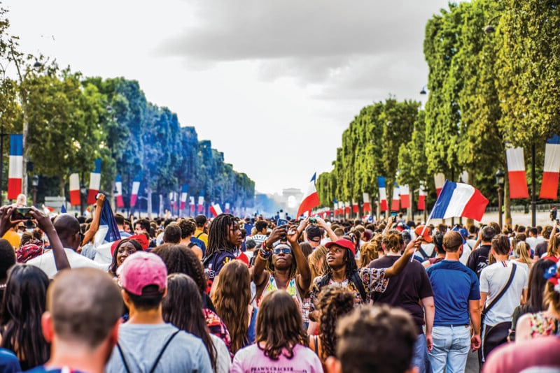 A large crowd gathers outdoors on a tree-lined street decorated with French flags, many people are taking photos and waving flags, with the Arc de Triomphe visible in the distance under a cloudy sky.