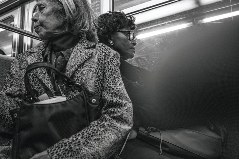 Two women sit side by side on public transit, facing different directions. One wears a leopard-print coat and headband, holding a handbag; the other wears glasses and dark clothing. The image is in black and white.