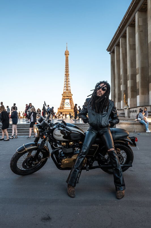 A person in black leather clothing poses confidently with a motorcycle in front of the Eiffel Tower in Paris, surrounded by people and clear blue sky.