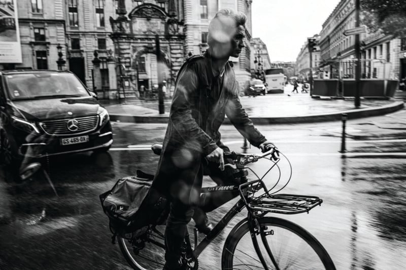 A person rides a bicycle on a wet city street, with raindrops visible on the camera lens. Cars and historic buildings appear in the background, creating a dynamic, urban scene in black and white.