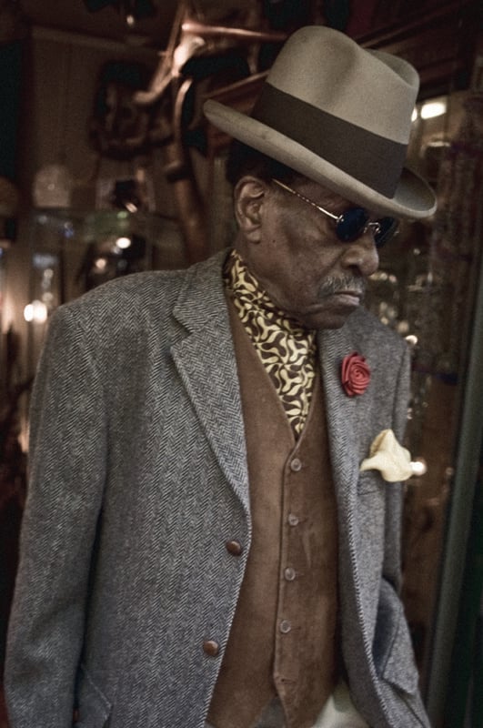 An older man wearing a grey herringbone blazer, brown vest, patterned scarf, sunglasses, and a fedora hat with a wide brim, accented with a red rose and yellow pocket square, stands in a dimly lit, stylish setting.