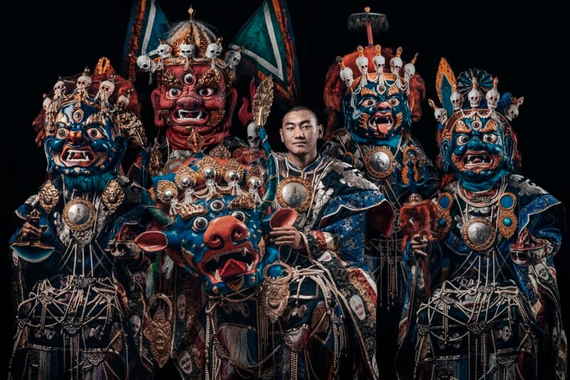 A group of five people wear ornate, colorful costumes and masks, including demonic faces with fangs, skulls, and horns, and elaborate jewelry, standing against a black background. One person in the center is unmasked.