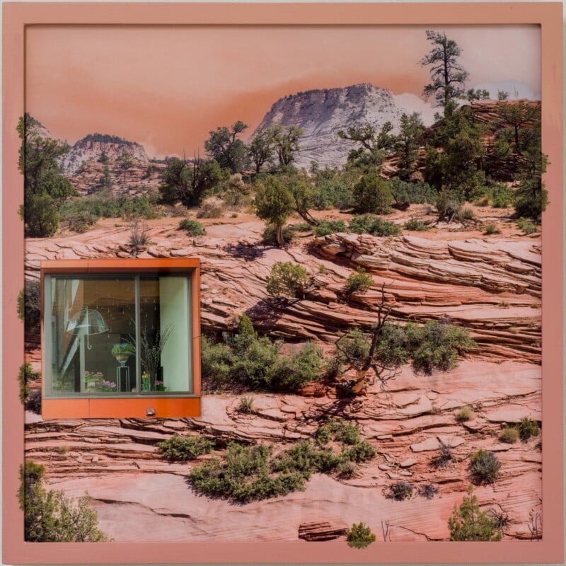 A framed scenic photograph of a rocky desert landscape with sparse greenery and distant mountains, featuring a cut-out window displaying indoor plants inside a modern room.