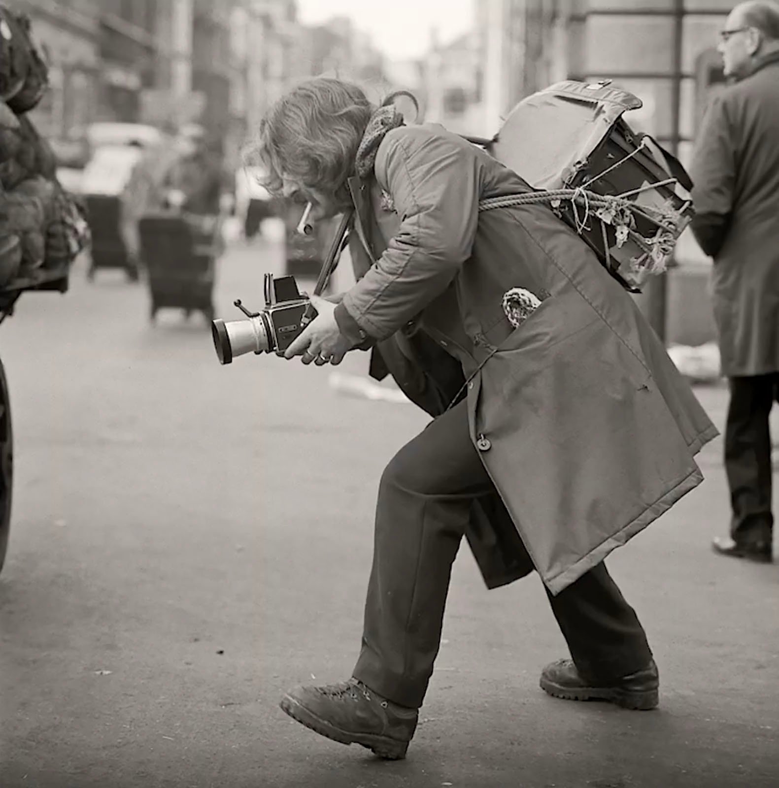 A person in a worn coat and backpack crouches on a city street, aiming a vintage camera. They appear focused on capturing a photo. Other people and blurred buildings are visible in the background.