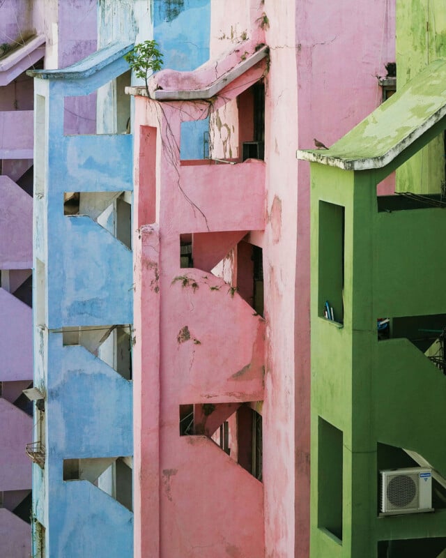 A close-up of a building with pastel blue, pink, and green painted walls, featuring geometric balconies, an air conditioner unit, and a small plant growing from a ledge. The paint is peeling in various areas.