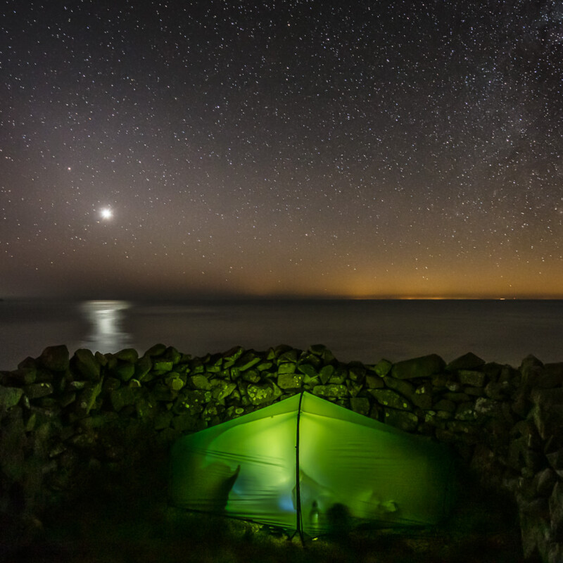 A glowing green tent sits behind a stone wall by calm water under a night sky filled with stars, with a bright planet or moon reflecting on the surface.