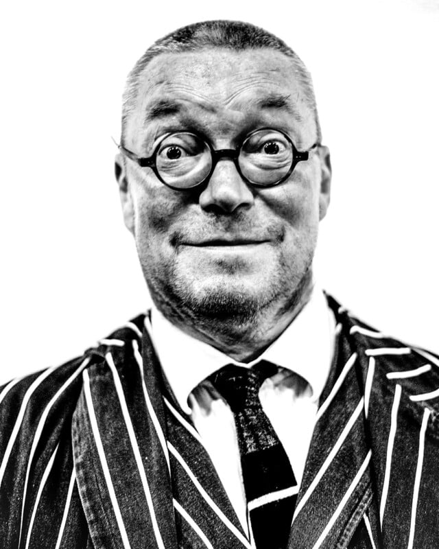 A man with round glasses and a short haircut smiles widely. He is wearing a pinstripe suit, tie, and white shirt. The black and white photo has a plain, bright background.