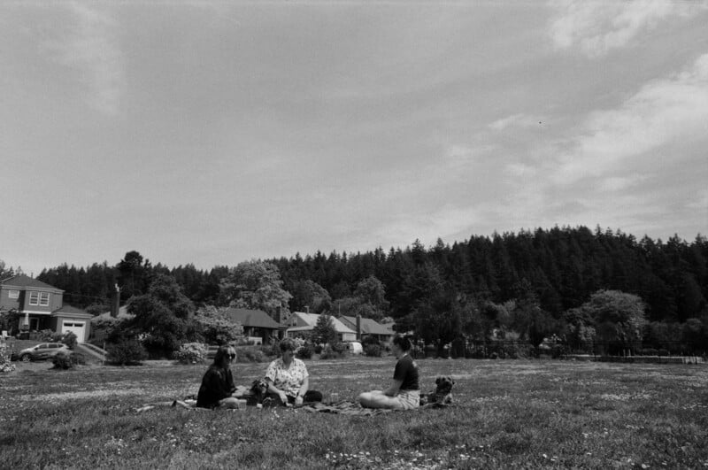 Three people sit on a blanket in an open grassy field, surrounded by houses and trees, with a forest in the background under a partly cloudy sky. The photo is in black and white.