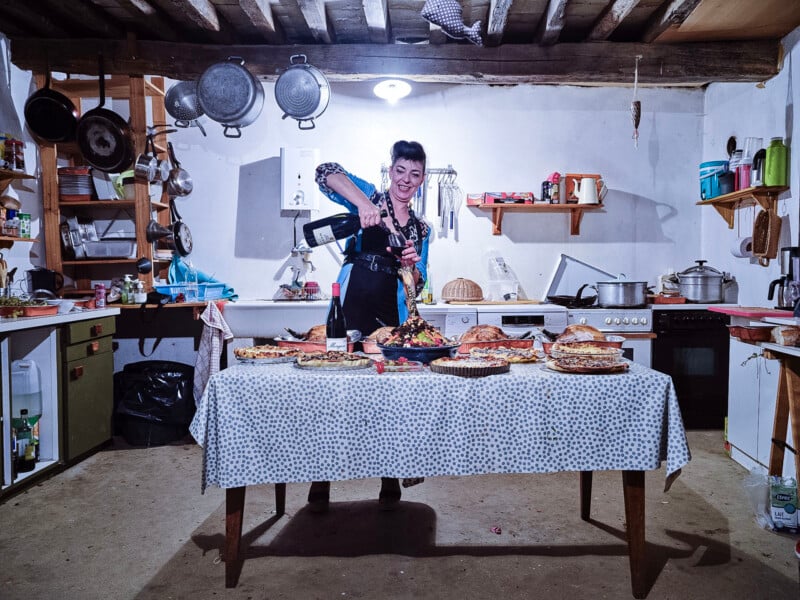 A woman in a rustic kitchen pours wine over a plate of food on a table covered with various dishes; pots, pans, and utensils hang from the wall and ceiling.
