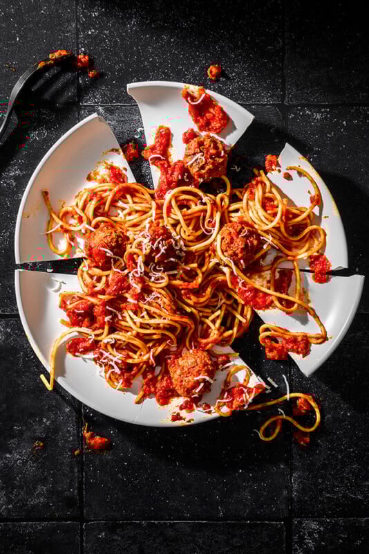 A plate of spaghetti with meatballs and tomato sauce sits on a black surface. The plate is broken into several pieces, with food and sauce spilled around the shards. A fork lies in the top left corner.