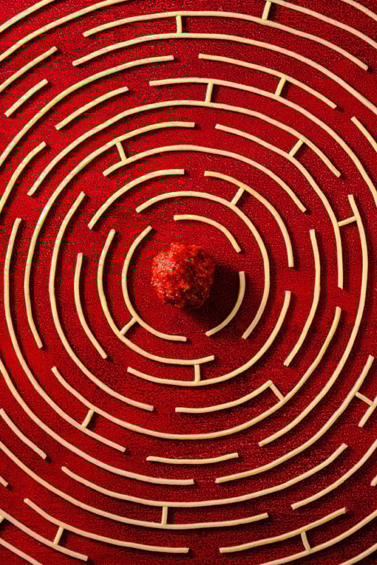 A circular maze with cream-colored walls on a red surface, viewed from above. At the center of the maze is a pile of red granules or a textured red sphere.