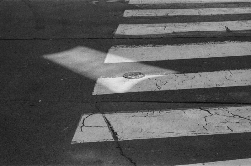 Black and white photo of a crosswalk with cracked lines and a round metal object on the pavement. A diagonal patch of sunlight highlights part of the crosswalk, contrasting with the surrounding shadows.