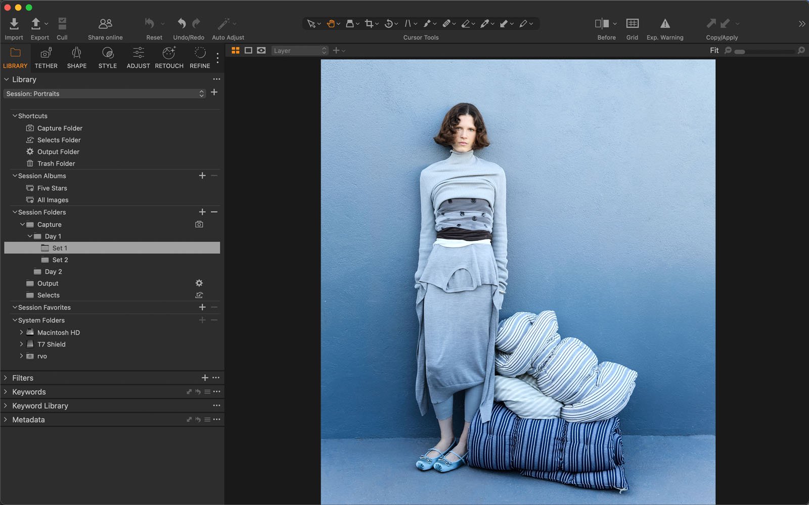 A woman in layered, monochromatic grey clothing stands against a blue wall next to a stack of blue-and-white striped pillows. The photo editing software interface is visible around the image.