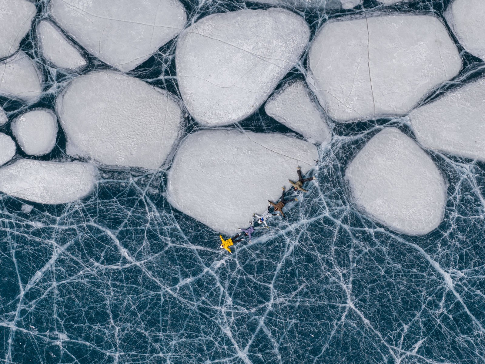 Aerial view of a dog sled team pulling a person across a frozen lake with large cracked ice sheets and intricate white fracture lines.