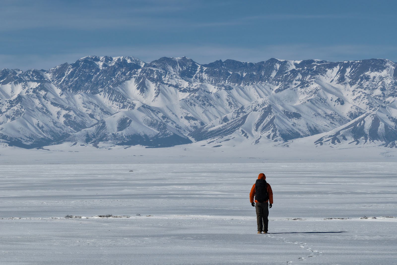 A person in an orange jacket stands alone on a vast snowy plain, facing a distant range of tall, snow-covered mountains under a blue sky.