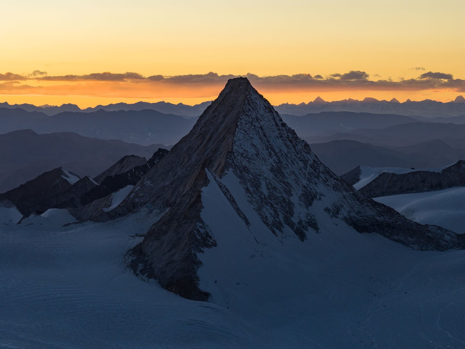 Snow-covered mountain peak silhouetted against an orange and yellow sunrise sky, with layers of distant mountain ranges visible on the horizon.