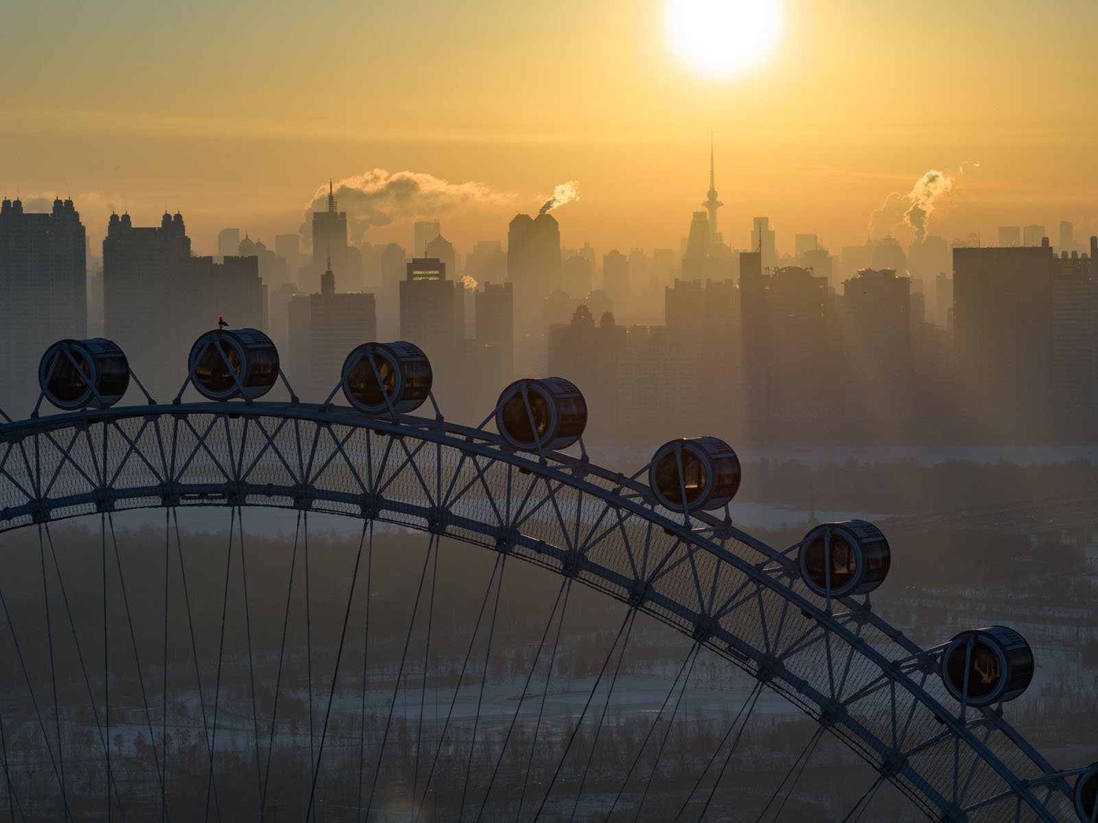 A large Ferris wheel in the foreground with a city skyline in the background at sunrise or sunset, with mist and sunlight creating a warm, golden atmosphere.