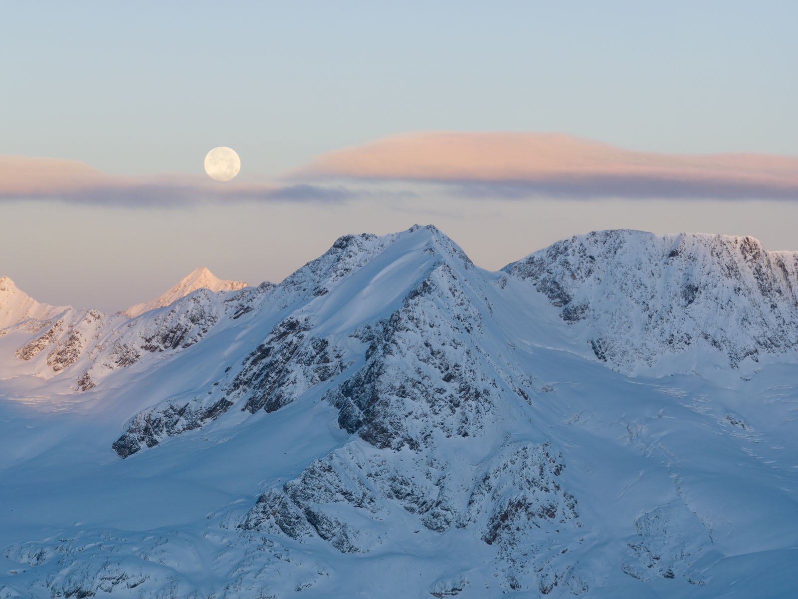 Snow-covered mountain peaks under a pale blue sky at dawn or dusk, with a full moon rising above the horizon and soft pink-tinged clouds drifting across the scene.