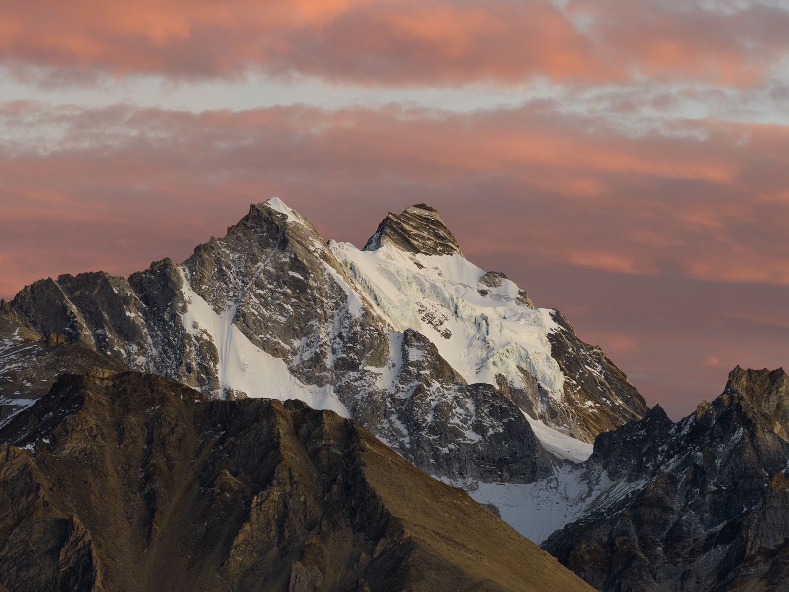 Snow-capped mountain peak under a dramatic sky with pink and orange clouds at sunset, with rugged, rocky slopes in the foreground.