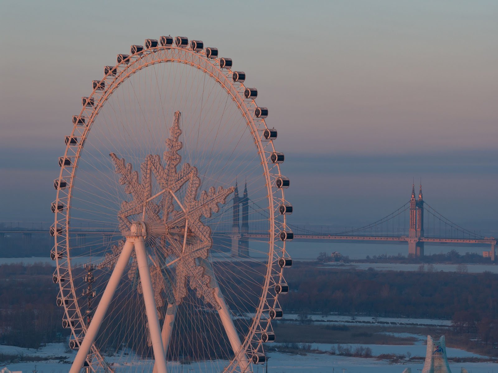 A large Ferris wheel with snowflake-shaped spokes stands in the foreground at sunset, with a suspension bridge and a river visible in the background. Snow covers the ground and soft pink light bathes the scene.