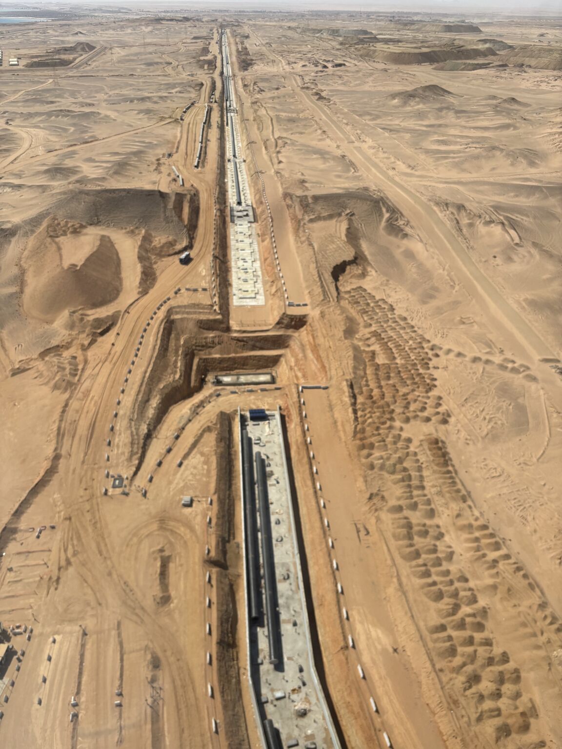 Aerial view of a large construction site in a desert, showing two long, parallel, rectangular concrete structures surrounded by sandy terrain, excavation work, and construction vehicles. The horizon is visible in the distance.
