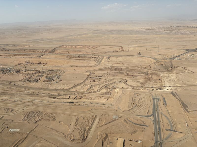Aerial view of a vast desert construction site with dirt roads, machinery, scattered structures, and cleared land under a hazy sky, surrounded by empty desert landscape.