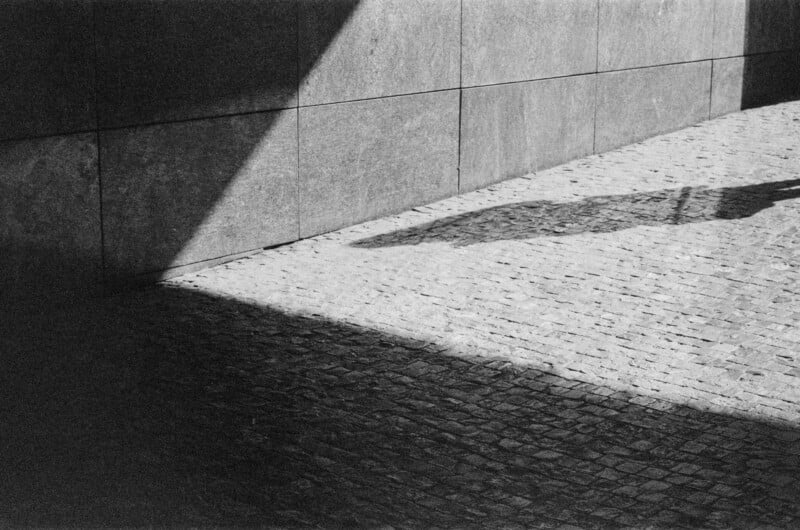 Black and white photo of a sunlit cobblestone street next to a stone wall, with geometric shadows and the partial shadow of a person holding something, possibly an umbrella, entering from the right.