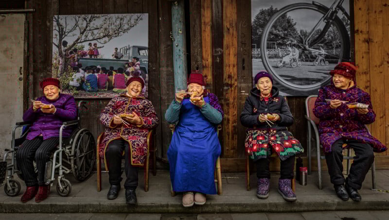 Five elderly women, wearing colorful clothing and hats, sit on chairs outside a wooden building, eating food and smiling. Two black-and-white photographs are displayed on the wall behind them.