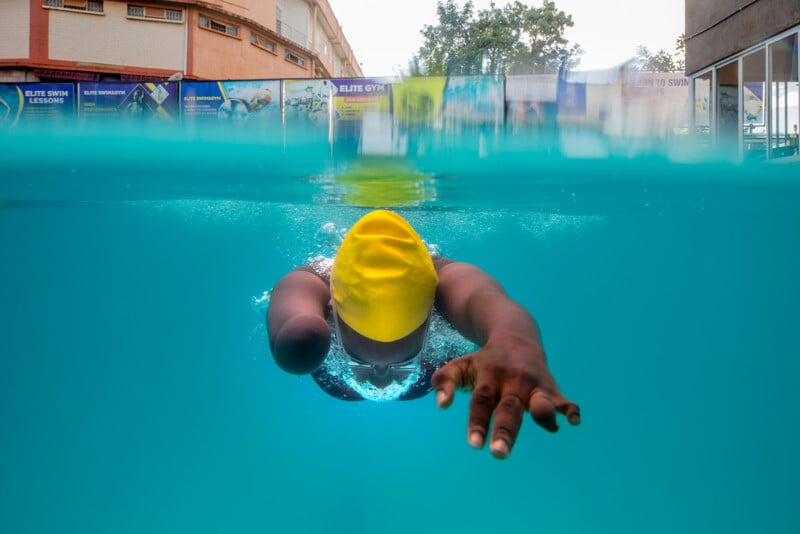 A swimmer wearing a yellow cap is captured mid-stroke underwater in a pool, reaching forward with one arm. The upper half of the image shows the poolside and banners outside.