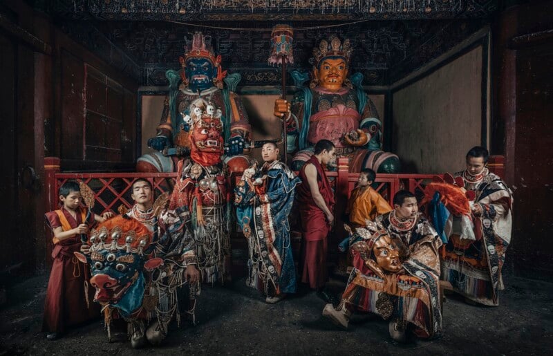 A group of monks and children dressed in ornate traditional costumes pose in front of large, colorful statues inside a dimly lit temple or shrine. The scene is rich in intricate details and vibrant patterns.