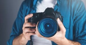 A person in a blue plaid shirt holds a professional DSLR camera, aiming the lens directly at the viewer. The camera lens is in sharp focus, while the background and person are blurred.