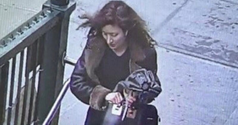 A woman with wavy brown hair and a dark coat walks on a sidewalk next to a stair railing, holding a bag and another item in her hands. The photo appears to be from a security camera.