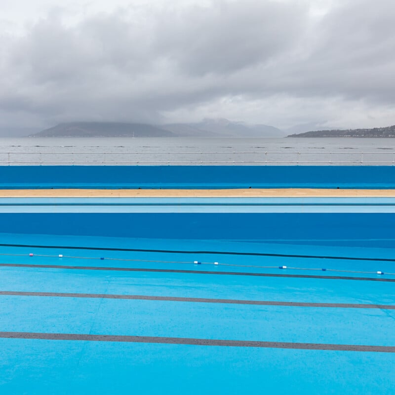 An empty outdoor swimming pool with blue lanes next to the sea, under a cloudy sky with mountains in the distance.