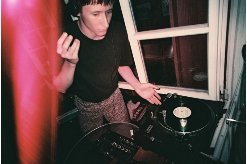A person wearing headphones stands by a turntable and mixer, appearing focused while DJing indoors. Pink light streaks across the left side of the image. There is a window in the background.