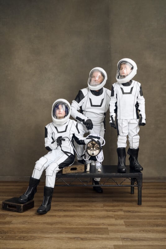 Three children wearing white and black space suits pose indoors with a small dog also dressed in a matching space suit. The children stand and sit on a bench against a plain brown background.