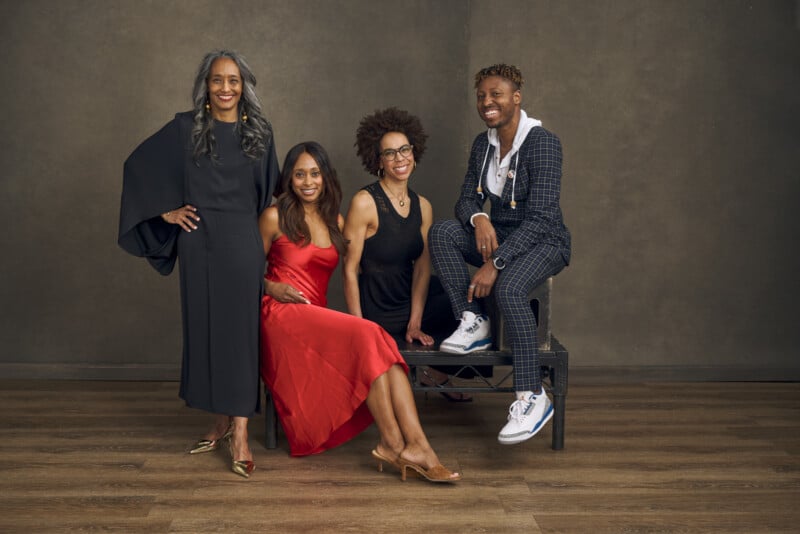 Four stylish people pose together in a studio: one stands in a black dress, one sits in a red dress, one in a black outfit kneels, and one sits on a bench in a plaid suit and sneakers, all smiling warmly.