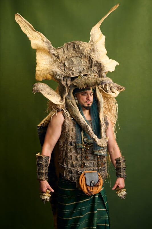 A person wearing an elaborate dragon-inspired costume with a large dragon headpiece, textured armor, and accessories, standing against a green background.