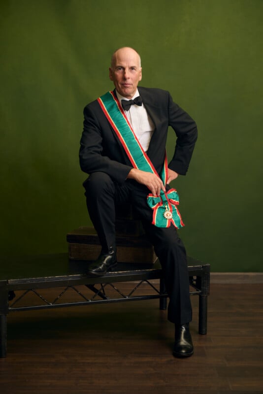 A man in a tuxedo and bow tie sits on a bench against a green backdrop, wearing a green, white, and red sash across his chest and holding a matching sash with a decorative medal.