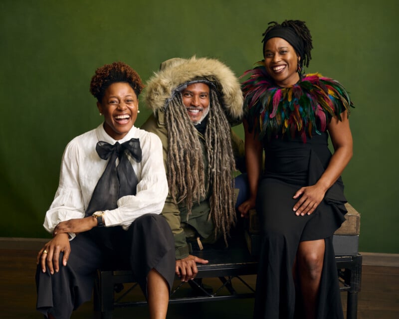 Three people sit together smiling against a green background. One wears a white shirt with a large black bow, another wears a hooded parka with long gray dreadlocks, and the third wears a black dress with a colorful feather collar.