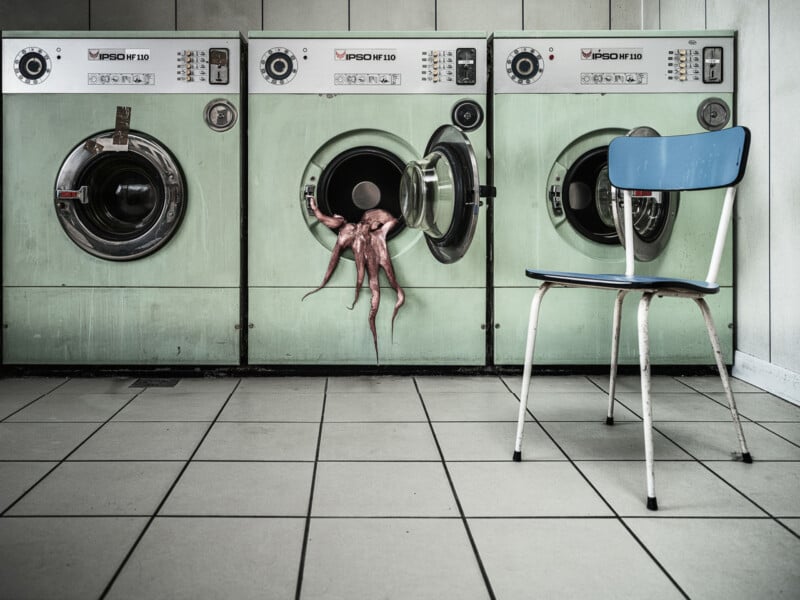 An octopus hangs partly out of an open washing machine in a laundromat with pale green washers and a blue chair on a tiled floor.
