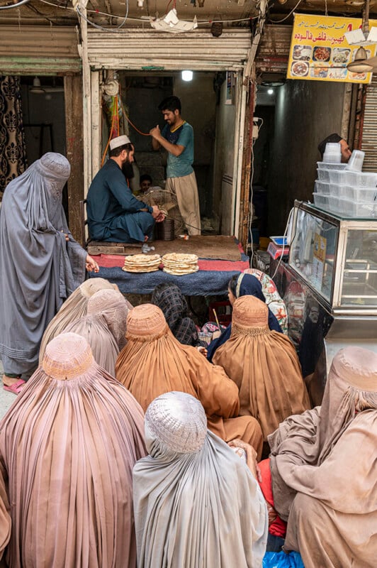 A group of women in burqas sit in front of a bakery stall, where two men are preparing and selling bread. The shop has naan displayed on a table and a sign with food photos above.