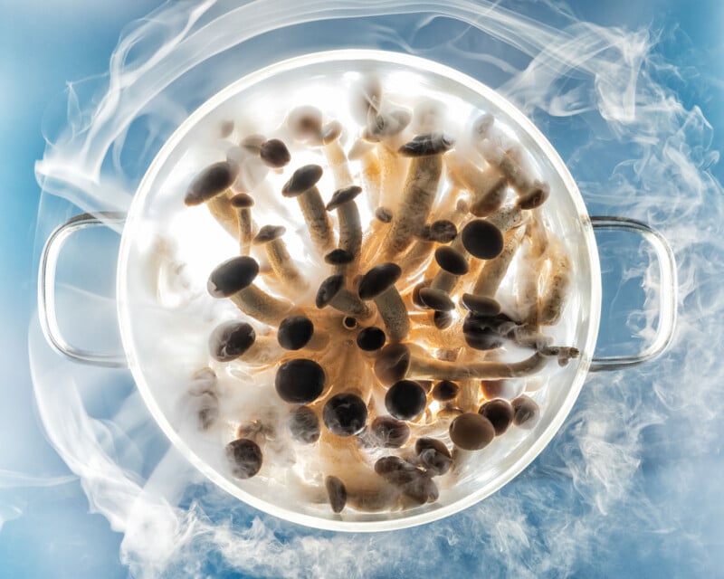 Overhead view of a pot filled with mushrooms, surrounded by swirling white smoke or steam against a blue background. The mushrooms are tightly packed and illuminated from above.