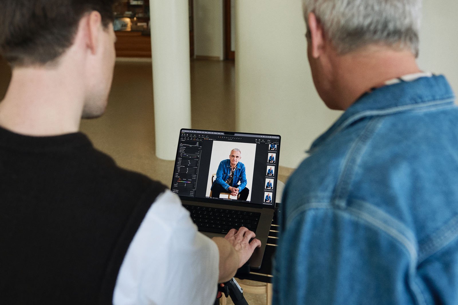 Two people look at a laptop screen displaying a portrait photo of an older man in a blue shirt, with photo editing software open showing adjustment tools and image thumbnails on the side.