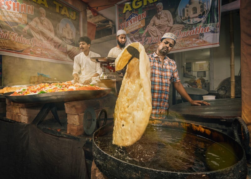 A man fries a large piece of bread in a deep pan at an outdoor food stall, while two others prepare colorful dishes beside him. Banners and vibrant food can be seen in the background.