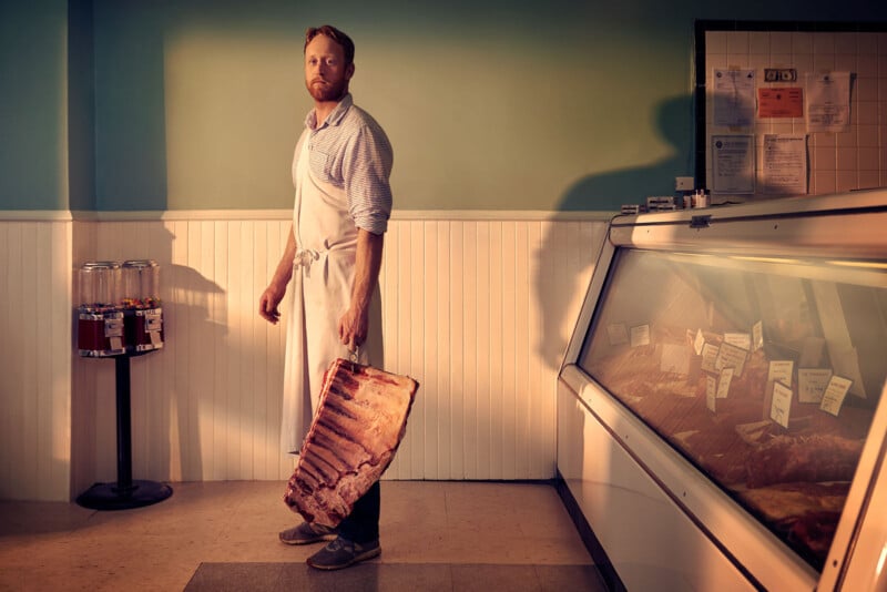 A butcher wearing an apron stands in a shop, holding a large rack of ribs. He faces forward, with a meat display case and labeled cuts to his right and candy dispensers on his left against a pale green and white wall.