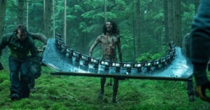 A person covered in mud stands in a forest while two crew members carry a large rig with multiple cameras mounted in a curved row; the scene appears to be from a film set.
