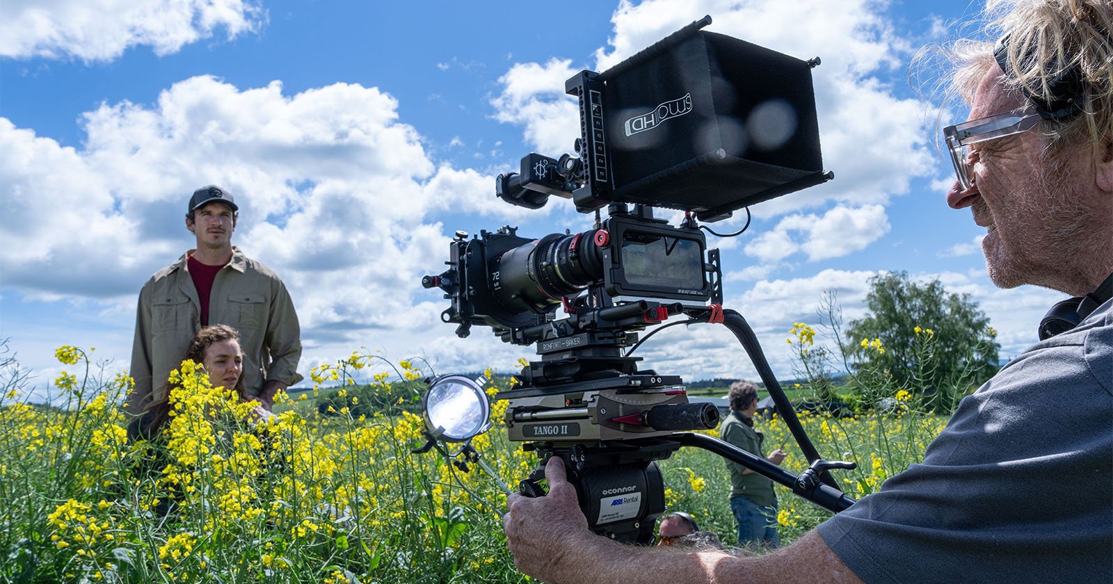 A film crew shoots a scene in a field of yellow flowers under a partly cloudy sky; one person operates a camera while two actors stand among the flowers.