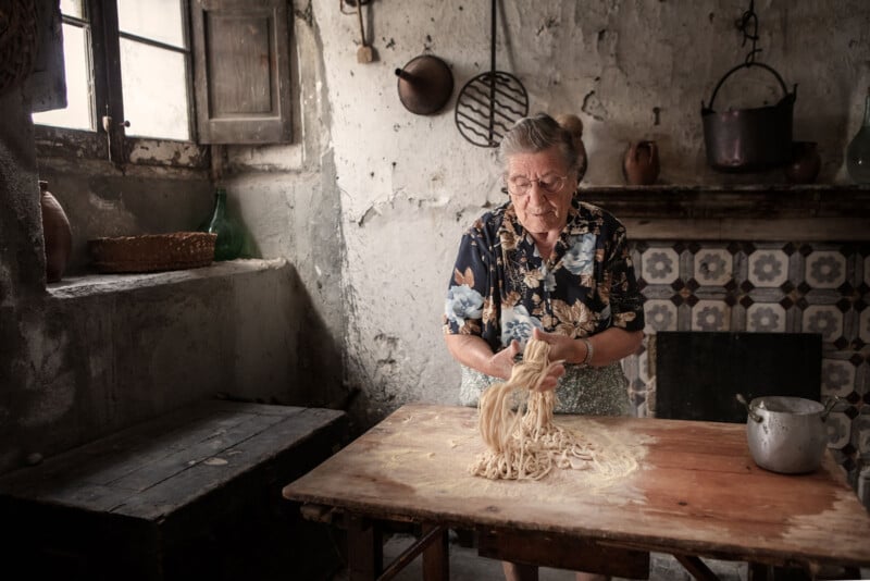 An elderly woman in a floral shirt makes fresh pasta on a wooden table in a rustic kitchen with worn walls, tiled fireplace, and vintage utensils. Light streams in from a window on the left.