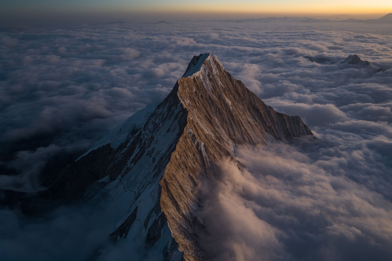 A dramatic mountain peak rises sharply above a sea of clouds at sunset, its rugged slopes partially covered in snow and bathed in warm golden light.