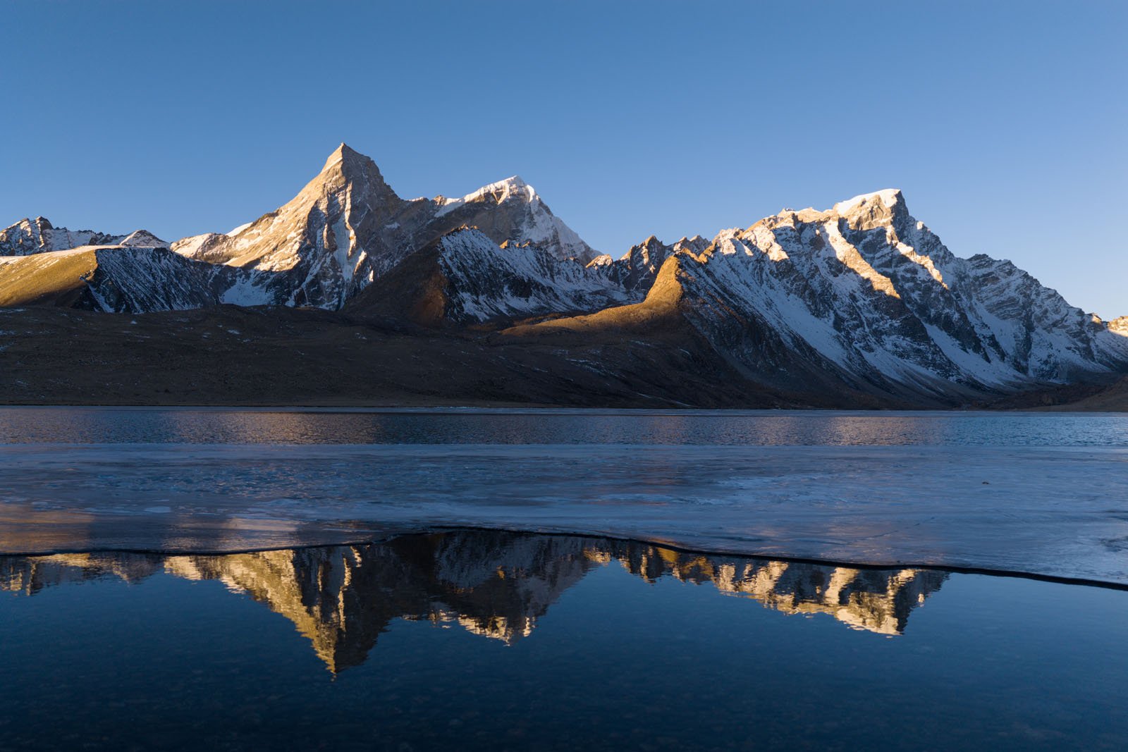Snow-capped mountain peaks are reflected in a calm, clear lake under a blue sky, with sunlight casting warm highlights on the rugged slopes and faint ice along the water’s edge.